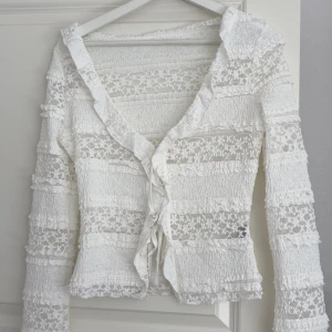 Vit spetsblus med knyt från SHEIN - Supersöt vit blus från SHEIN i storlek XS. Blusen har långa ärmar, v-ringning och knyt framtill. Den är gjord i spets med volangdetaljer och har en romantisk vibe. Perfekt att styla med jeans eller kjol för en trendig look.