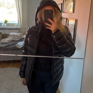 Svart pufferjacka från Colmar - Säljer en svart pufferjacka från Colmar med glansig finish och huva. Jackan har en tydlig logga på ärmen och dragkedja framtill. Fodret har tryckt Colmar Originals-mönster. Passformen är normal och jackan är perfekt för kalla dagar.