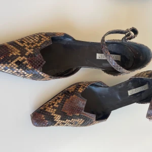 Bruna pumps i ormskinn från Footlight - Unika pumps från Footlight i äkta ormskinn med brunt och beige mönster. Skorna har spetsig tå, låg klack och justerbar rem runt vristen. Tillverkade i Italien med lädersula. Perfekta för dig som vill sticka ut med ett djurmönstrat statement. Sulan på vänsterskon behöver limmas fast längst fram. Annars är skicket bra!