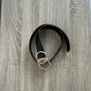 Svart bälte med dubbla guldringar - Snyggt svart bälte med två stora, runda guldfärgade ringar som spänne. Bältet är i slät skinnimitation och har en stilren, modern look som passar till jeans eller klänning. Perfekt accessoar för att lyfta din outfit med en cool detalj.