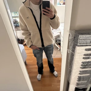 Beige fleecejacka  - Säljer en beige fleecejacka med svart dragkedja och hög krage. Jackan har en clean look med diskret Lacoste-logga på bröstet och två sidofickor. Perfekt för dig som gillar chill och stilren vibe.
