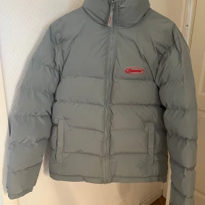 Grå pufferjacka från Trapstar - Säljer en grå pufferjacka från Trapstar med fet röd logga broderad på ryggen och mindre logga på bröstet. Jackan har hög krage, två fickor fram och dragkedja med Trapstar-detalj. Perfekt för kalla dagar och riktigt clean stil.