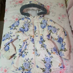 Blommig pufferjacka med huva - Säljer en fluffig pufferjacka med blommigt mönster i blått och ljusorange på vit bakgrund. Jackan har en mysig grå fuskpäls runt huvan, dragkedja framtill och två fickor. Midjan är lätt markerad för snygg passform. Perfekt för kalla dagar!