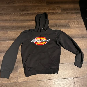 Svart Dickies hoodie med logga - Svart hoodie från Dickies i storlek small med stor färgglad Dickies-logga på bröstet. Klassisk känguruficka framtill, huva med snörning och ribbade muddar. Perfekt för dig som gillar streetwear och vill ha en skön och stilren tröja.