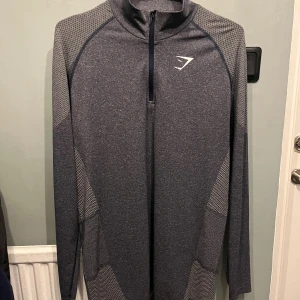 Mörkgrå Gymshark träningströja half zip - Sportig mörkgrå långärmad träningströja från Gymshark med half zip och diskret mönstrade partier på ärmar och sidor. Tillverkad i en blandning av polyester och nylon för en skön och stretchig känsla. Perfekt för gymmet eller löprundan.