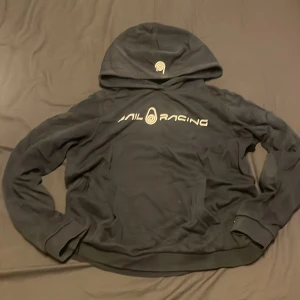 Svart Sail Racing hoodie med tryck - Svart hoodie från Sail Racing med vit logga och text på bröstet samt liten logga på huvan. Klassisk känguruficka framtill, ribbade muddar och mjukt material. Perfekt för dig som gillar sportig och clean stil.