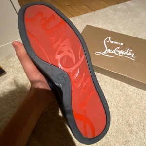 Christian Louboutin svarta sneakers med nitar - Svarta sneakers från Christian Louboutin med ikonisk röd sula och coola svarta nitar på tån. Skorna har snörning och är tillverkade i mocka med platt sula. Perfekt för dig som vill sticka ut med en edgy och lyxig stil.