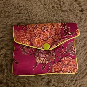 Blommig kuvertväska i rosa och gult - Kuvertväska i glansigt tyg med stora blommor i orange, gult och rosa på en starkt rosa bakgrund. Väskan har gul kant, tryckknapp och innerfack med dragkedja. Perfekt för att ge din outfit en färgklick.