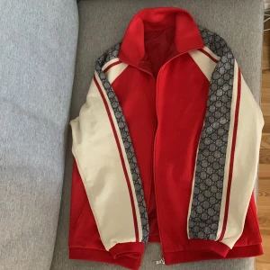 Gucci röd och vit bomberjacka - Säljer en snygg Gucci bomberjacka i rött och vitt med klassiska GG-mönstrade paneler längs ärmarna. Jackan har dragkedja framtill, ribbade muddar och sportig krage. Materialet är polyester och jackan har en loose passform. Perfekt för dig som vill sticka ut med lyxig streetstyle. Lappen i nacken saknas. 