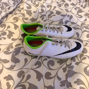 Nike Mercurial vita fotbollsskor - Nike Mercurial fotbollsskor i vitt med svarta detaljer och limegröna insidor samt sulor. Skorna har snörning och platt sula med dobbar för konstgräs. Snygg, sportig design med stor Nike-logga på sidan och tå. Perfekt för dig som vill sticka ut på planen.