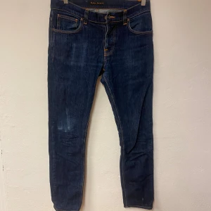 Mörkblå jeans från Nudie Jeans Co W32 L36 - Snygga mörkblå jeans från Nudie Jeans Co med klassisk femficksdesign och orange kontrastsömmar. Modellen har rak passform och är tillverkad i slitstarkt bomullsjeansmaterial. Bakfickorna har Nudie Jeans logga och detaljer i orange.