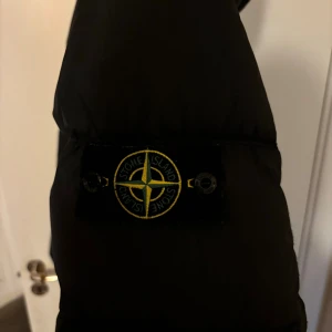 Svart pufferjacka från Stone Island - Svart pufferjacka från Stone Island med ikonisk patch på ärmen och huva. Jackan har två stora fickor med knappar framtill och dragkedja. Materialet är syntet och dun, perfekt för kalla dagar. Snygg och stilren design med klassiska detaljer.