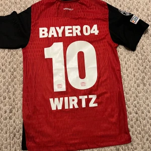 Bayer Leverkusen Wirtz matchtröja - Säljer en officiell Bayer Leverkusen fotbollströja från Castore med Wirtz och nummer 10 på ryggen. Tröjan är röd med svarta ärmar, klubbmärke och sponsortryck framtill samt Champions League-märke på ärmen. Tillverkad i lätt och ventilerande material.