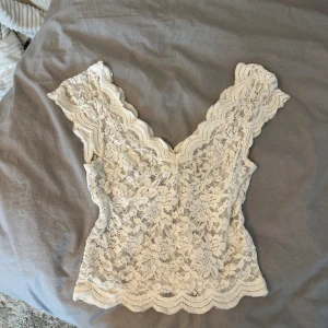Vit spetsig linnetopp från Stradivarius - Säljer en superfin vit linnetopp från Stradivarius i storlek XS. Toppen är helt i spets med blommigt mönster och har en snygg v-ringad form både fram och bak. Ärmlös design med vågiga kanter som ger en romantisk vibe. Perfekt att styla med jeans eller kjol!