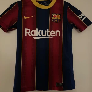 Barcelona fotbollströja Nike M - Säljer en officiell FC Barcelona fotbollströja från Nike i storlek M. Tröjan har klassiska blå och röda vertikala ränder med gula detaljer, klubbmärke på bröstet och Rakuten-tryck. Kortärmad och tillverkad i lätt polyester för bästa komfort på planen.