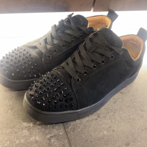 Säljer Svarta sneakers med nitar - Säljer Svarta sneakers i mocka med coola svarta nitar på tån. Skorna har snörning och en platt sula, samt en stilren och edgy look. Insidan är beige och de har en klassisk rund tå. Perfekta för dig som vill sticka ut med din stil.