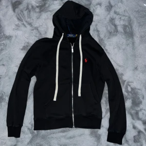 Polo Ralph Lauren zip hoodie - Svart hoodie från Polo Ralph Lauren med vit snörning och dragkedja framtill. Tröjan har huva, två fickor och rött broderat Polo-logo på bröstet. Insidan är mjuk och fleecefodrad för extra komfort. Perfekt för chill dagar och streetwear.
