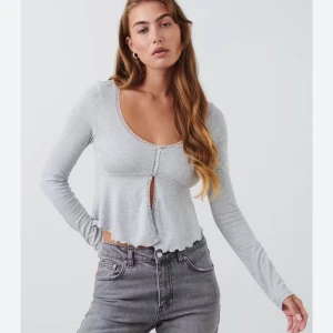 Grå croppad blus med spetsdetaljer - Superfin ljusgrå croppad blus med lång ärm och spetskant längs halsringningen och framtill. Toppen har en liten rosett och knäppning framtill, samt vågig kant nedtill för extra cute vibe. Perfekt att styla med jeans eller kjol för en chill look. Från Gina tricot i storelk S