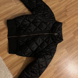 Svart quiltad bomberjacka Vera & Lucy - Quiltad svart bomberjacka från Vera & Lucy med orange foder och snygg dragkedja framtill. Jackan har ribbade muddar och en lite puffig look som passar perfekt till streetwear. Klassisk bombermodell med krage och stilren design.