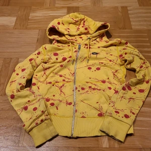Gul hoodie med rött splatter från Nike - Nike Sportswear hoodie i gul färg med unikt rött splattermönster. Tröjan har huva med snörning, hel dragkedja framtill och ribbade muddar. Perfekt för dig som gillar streetstyle och vill sticka ut med färg. Materialet är mjuk bomull och polyester.