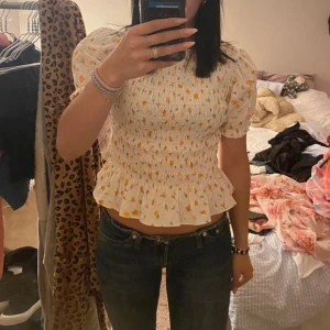 Apelsin blus från H&M Divided - Supersöt apelsin blus från H&M Divided i storlek S. Blusen är vit med små orange apelsiner på, har puffiga korta ärmar och smock över hela kroppen. Volangkant nedtill ger en extra girly vibe. Perfekt för att lyfta din sommarstil! Köpt på plick