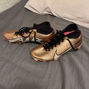 Nike Mercurial fotbollsskor guld/svart - Säljer ett par Nike Mercurial fotbollsskor i metallic guld med svarta detaljer och snörning. Skorna har högre strumpliknande skaft, rosa innersula och vita samt rosa dobbar. Perfekta för dig som vill sticka ut på planen!