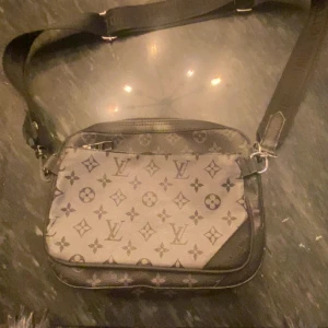 Louis Vuitton axelväska monogram - Snygg axelväska från Louis Vuitton med klassiskt monogrammönster i svart och grå. Väskan har justerbar axelrem, två fack med dragkedja och detaljer i skinn. Perfekt storlek för det viktigaste och ikonisk design med LV-logga och blommor.