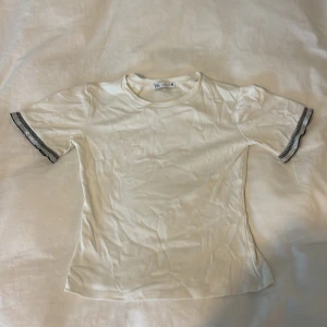 Vit kortärmad topp från Zara - En vit topp från Zara med rund halsringning och korta ärmar. Ärmsluten har en svart och silverfärgad ribbad detalj