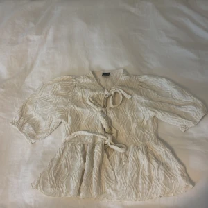 Beige omlottblus med knytband - beige omlottblus med knytband i midjan och vid halsen. Blusen har en lätt skrynklig struktur, trekvartsärmar och en luftig passform