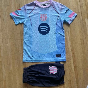 Barça Lamine Yamal tröja & shorts - Säljer en FC Barcelona matchtröja med Lamine Yamal #10 och tillhörande shorts. Tröjan är ljusblå med rosa och mörkblå detaljer, kortärmad och har tryck från Spotify och UNHCR. Materialet är lätt och andas, perfekt för fotboll. Shorts i mörkblått med Barça-emblem.