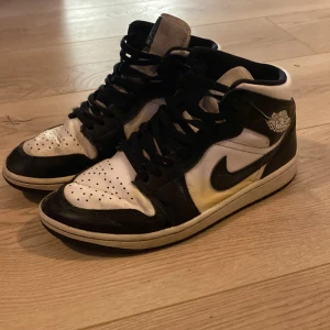 Nike Air Jordan 1 svartvita sneakers - Svartvita Nike Air Jordan 1 sneakers med klassisk hög siluett. Skorna har svart snörning, perforerad tå och ikoniska Air Jordan-loggan på sidan. Tillverkade i skinn med vit bas och svarta detaljer. Perfekta för dig som gillar streetstyle och vill sticka ut.