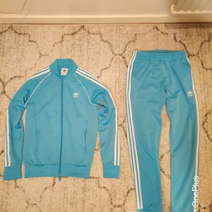 Adidas blå träningsdress S - Säljer en klassisk Adidas träningsdress i ljusblått med vita ränder längs ärmar och ben. Setet består av zip-jacka och byxa, båda med Adidas logga framtill. Jackan har hög krage och dragkedja, byxan har resår i midjan. Perfekt för dig som gillar retrostil och sportiga vibes.
