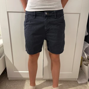 Mörkblå shorts från Nudie Jeans - Säljer ett par mörkblå shorts från Nudie Jeans med klassisk passform. De har fickor fram och bak, bälteshällor och stängs med knapp och dragkedja. Materialet är mjuk bomull som känns skönt mot huden. Perfekta för varma dagar och enkel stil. Finns en liten defekt i tyget baktill, se bild 2. 