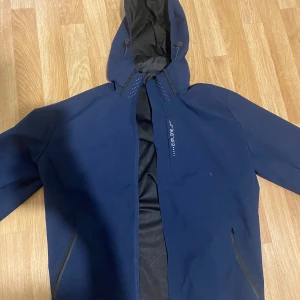 Blå vindjacka Explore med huva - Snygg marinblå vindjacka från Explore med huva och svarta meshdetaljer på insidan. Jackan har dragkedjor på fickorna och en stilren design med tryckt logga på bröstet. Perfekt för blåsiga dagar och outdoor-aktiviteter.