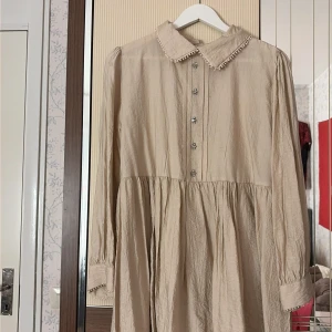 Beige långklänning från naqShe store - Långklänning i beige från naqShe store med dekorativa kristallknappar framtill, klassisk krage med söt kant och långa ärmar. Klänningen har en lös passform och är tillverkad i ett lätt, skrynkligt tyg som ger en avslappnad känsla.