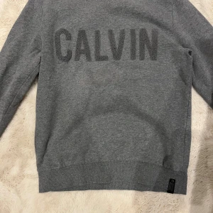 Grå sweatshirt från Calvin Klein Jeans - Snygg grå sweatshirt från Calvin Klein Jeans med stort CALVIN-text i frotté på bröstet. Tröjan har rund hals, ribbade muddar och långärmad passform. Perfekt för dig som gillar stilrena och klassiska plagg med en modern touch.