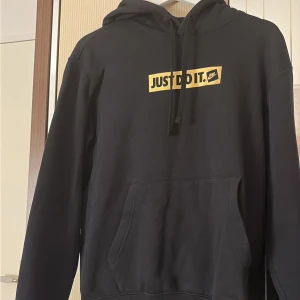 Svart Nike hoodie Just Do It S - Svart hoodie från Nike i storlek S med klassisk 'Just Do It.'-print i gult och svart på bröstet. Tröjan har huva med snörning och en stor magficka framtill. Perfekt för dig som gillar sportig och avslappnad stil.