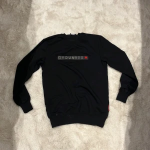 Svart Dsquared2 sweatshirt med tryck - Svart sweatshirt från Dsquared2 med silverfärgat DSQUARED-tryck och röd lönnlöv-detalj på bröstet. Tröjan har rund hals, ribbade muddar och är tillverkad i mjuk bomull. Perfekt för dig som gillar stilrena designer med en touch av street.