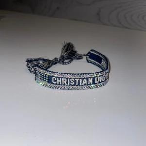 Glittrigt armband Christian Dior - Supersnyggt armband från Christian Dior med texten 'CHRISTIAN DIOR J'ADIOR' broderad i vitt på mörkblå bakgrund. Armbandet har glittriga detaljer och avslutas med stiliga tofsar i matchande färger. Perfekt statement piece för dig som gillar lyxiga accessoarer. Har massor med färger tillgängligt👍