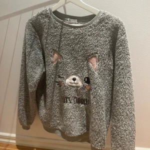 Grå fluffig tröja - Supermjuk och fluffig grå fleece-tröja med ett broderat djuransikte och texten 'Let's cuddle' på framsidan. Tröjan har långa ärmar och ribbad kant vid halsen. Perfekt för chilliga dagar när du vill mysa till max.