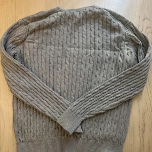 Grå stickad tröja från GANT - Snygg grå kabelstickad tröja från GANT med rund hals och broderad logga på bröstet. Tröjan har ribbade muddar vid ärmslut och nederkant, samt långärmad passform. Perfekt för dig som gillar klassisk stil med en modern touch.