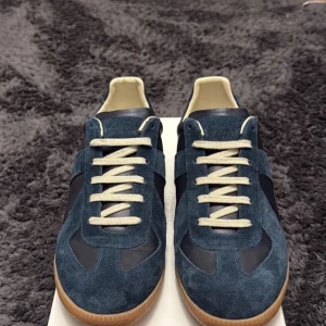 Maison Margiela blå sneakers i mocka - Maison Margiela sneakers i mörkblå mocka och skinn med beige snörning och gummisula. Klassisk rund tå och platt sula, stilren design med retrovibbar. Kommer med originalkartong och dustbags. Perfekt för dig som gillar exklusiva och snygga sneakers.