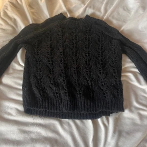 Svart stickad tröja med hålmönster - Svart stickad tröja med snyggt hålmönster och ribbade muddar vid ärmslut och nederkant. Tröjan har lång ärm och en klassisk rund halsringning. Perfekt för lager-på-lager och passar till många olika stilar.