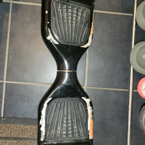 Hoverboard - Svart hoverboard med tydliga tecken på slitage och repor runt fotplattorna. Vissa delar av plasten är skadade och färgen har lossnat, men pedalerna ser ut att vara intakta. Perfekt för dig som vill ha ett prisvärt transportmedel och inte bryr dig om kosmetiska defekter.