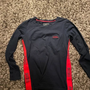 Mörkblå långärmad tröja från Diesel - Snygg mörkblå långärmad tröja från Diesel med röda detaljer längs sidorna där det står 'DIESEL' tryckt. Tröjan har rund halsringning och är tillverkad i mjuk bomull, perfekt för en avslappnad streetstyle-look.