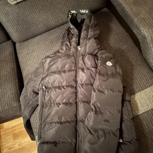 Svart Moncler pufferjacka med huva - Svart pufferjacka från Moncler med huva och dragkedja. Jackan har logotyp på ärmen, broderad Moncler-text på insidan av huvan och två fickor med dragkedja. Materialet är glansig polyester och jackan är vadderad för extra värme.