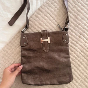 Brun axelväska med metallspänne - Snygg brun axelväska i skinn med justerbar rem och silverfärgat H-spänne framtill. Väskan har en praktisk dragkedja på baksidan och ett mönstrat innerfoder. Perfekt storlek för mobil, plånbok och småsaker. Stilren design med metallnitar och detaljer.