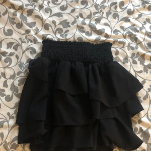 Svart volangkjol från H&M - Säljer en svart kortkjol från H&M med bred smock i midjan och flera lager volanger. Kjolen har en loose passform och är tillverkad i ett mjukt syntetmaterial som ger ett snyggt fall. Perfekt för dig som gillar volanger och vill ha en enkel men trendig look.🤍