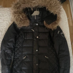 Rock and blue jacka M - Svart pufferjacka från rock and blue med stor fluffig päls på huvan. Jackan har quiltade detaljer, flera dragkedjor och en badge på ärmen. Snygg och varm modell med lång ärm och figurnära passform. Perfekt för vintern. Knappt använd. Nypris 3800kr, säljer för 450kr. 