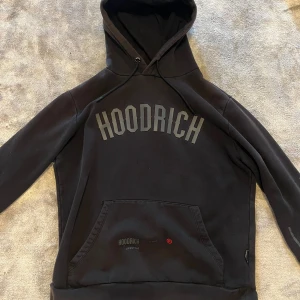 Svart hoodie från Hoodrich - Svart hoodie från Hoodrich med stor logga över bröstet och mindre tryck på fickan. Tröjan har huva med snörning och en klassisk magficka. Perfekt för dig som gillar streetwear och vill ha en snygg och bekväm look.
